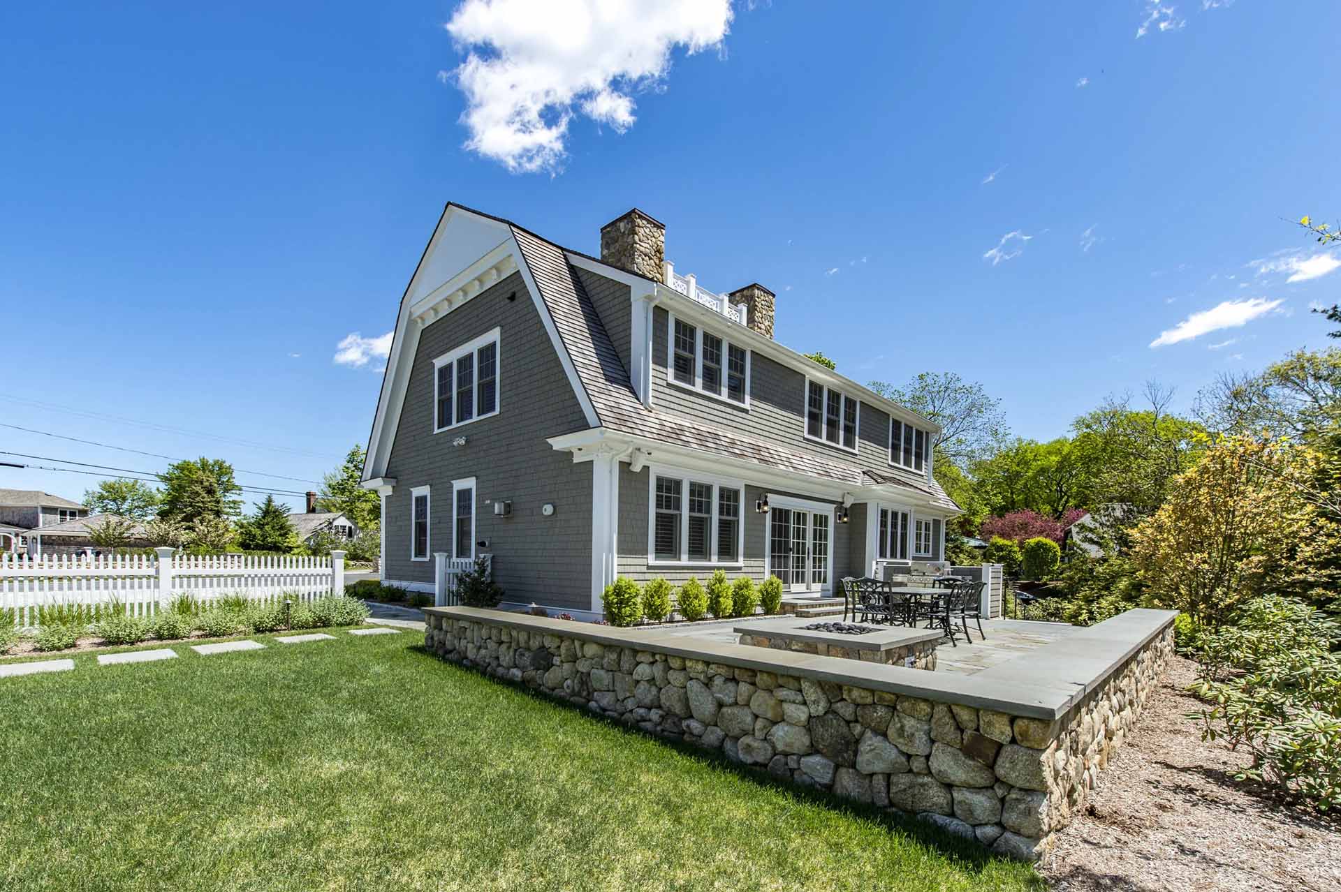 The rear façade, with a bluestone patio, stone retaining wall, and fire pit set against the home's classic gambrel roofline.