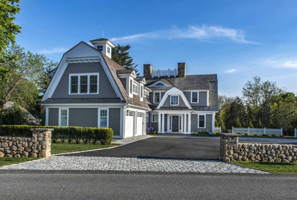 Coastal Home's front façade — grey shingle siding, twin stone chimneys, and a columned entry portico framed by a cobblestone motor court.