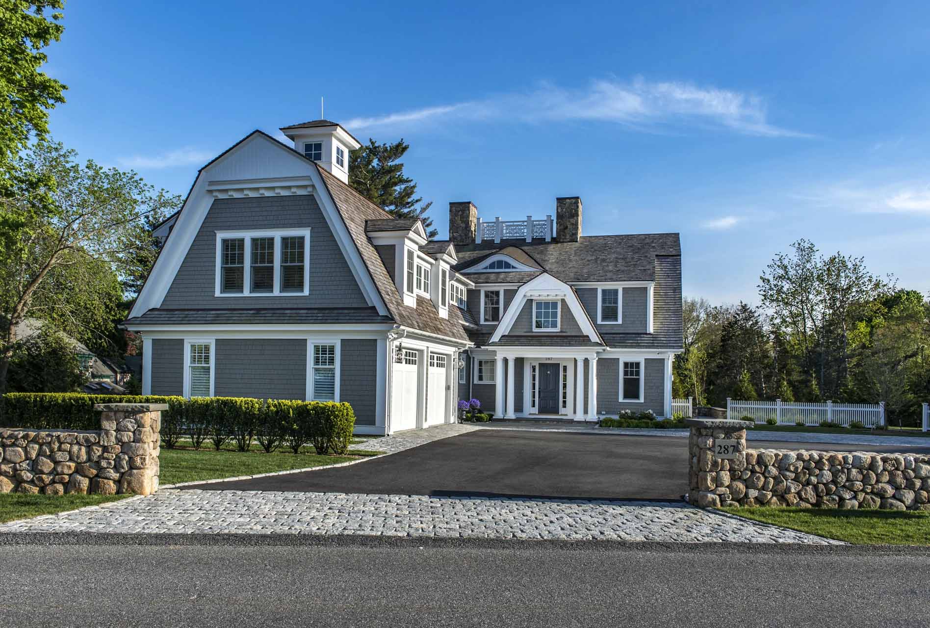 Coastal Home's front façade — grey shingle siding, twin stone chimneys, and a columned entry portico framed by a cobblestone motor court.